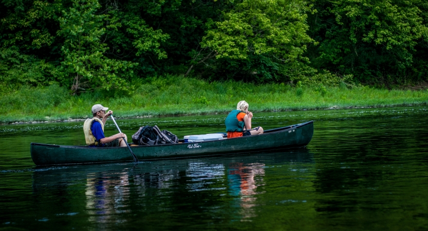 Two people wearing life jackets paddle a canoe on still water. On the shore behind them there are green trees. 